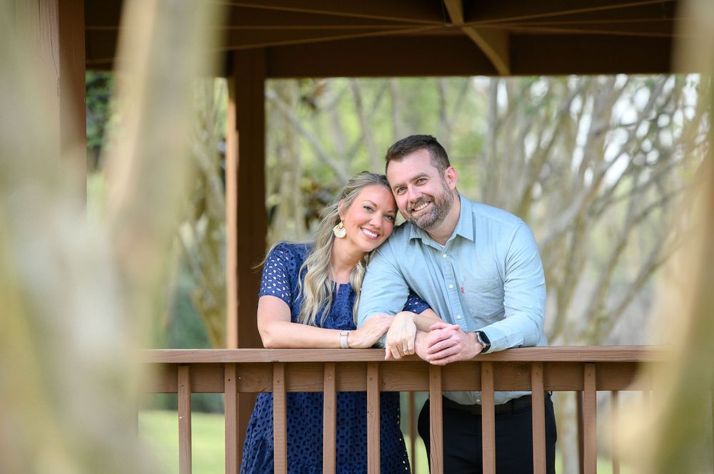 Happy couple smiling and leaning on wooden gazebo railing surrounded by lush green trees outdoors.