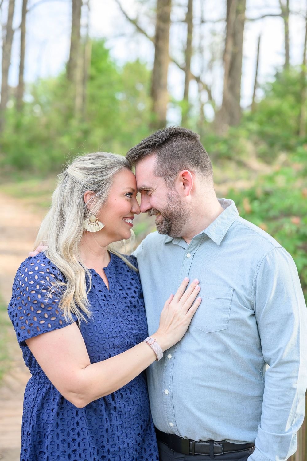 A smiling couple shares a romantic forehead touch outdoors, woman in blue lace dress, man in light blue shirt, forest path behind them.
