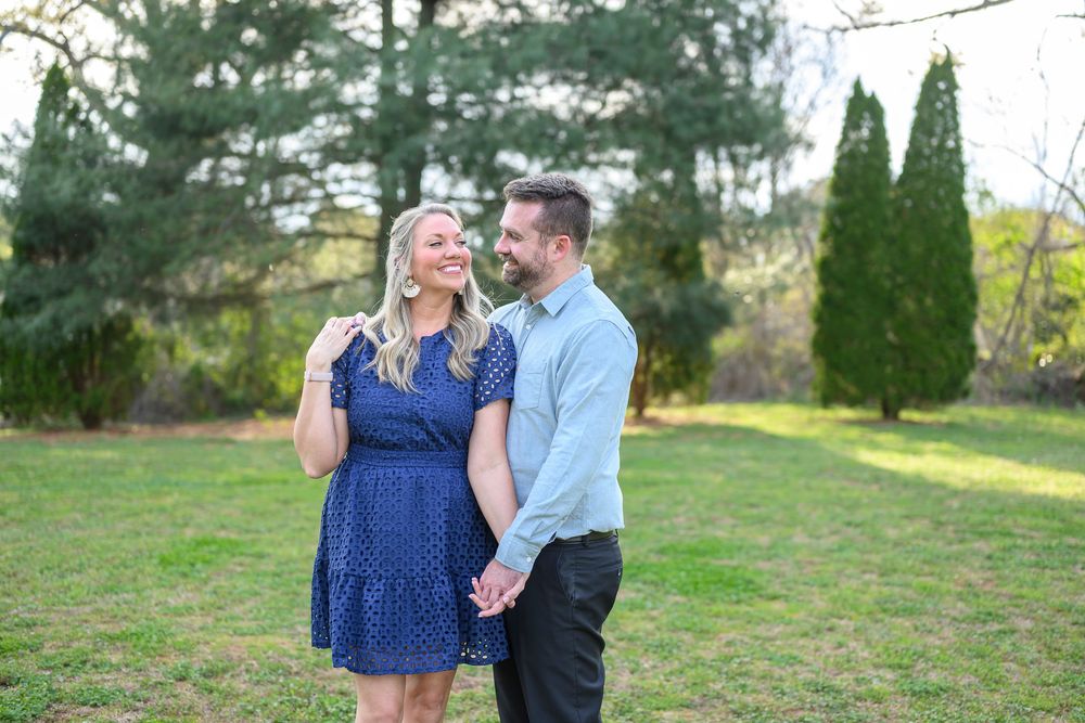 Happy couple holding hands and smiling in a lush green garden, woman in navy dress and man in light blue shirt.
