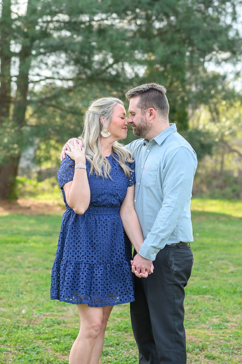 Romantic couple holding hands and touching foreheads in a sunny green park, woman in navy dress and man in light blue shirt.