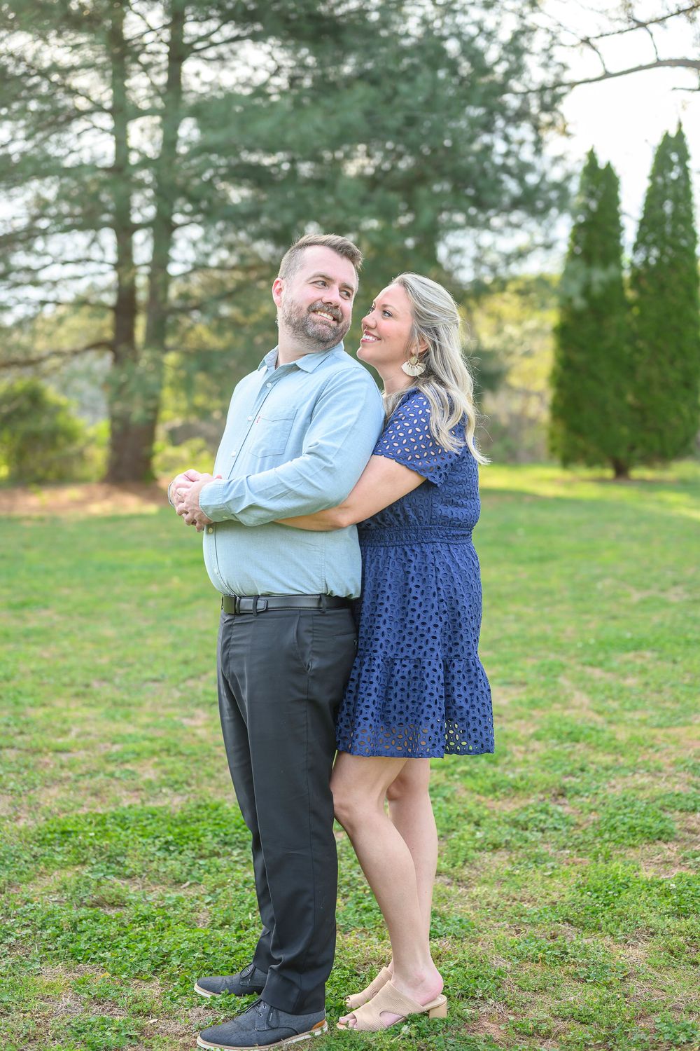 Couple embracing outdoors, woman in navy dress hugging man in light blue shirt, smiling in a lush green park setting.