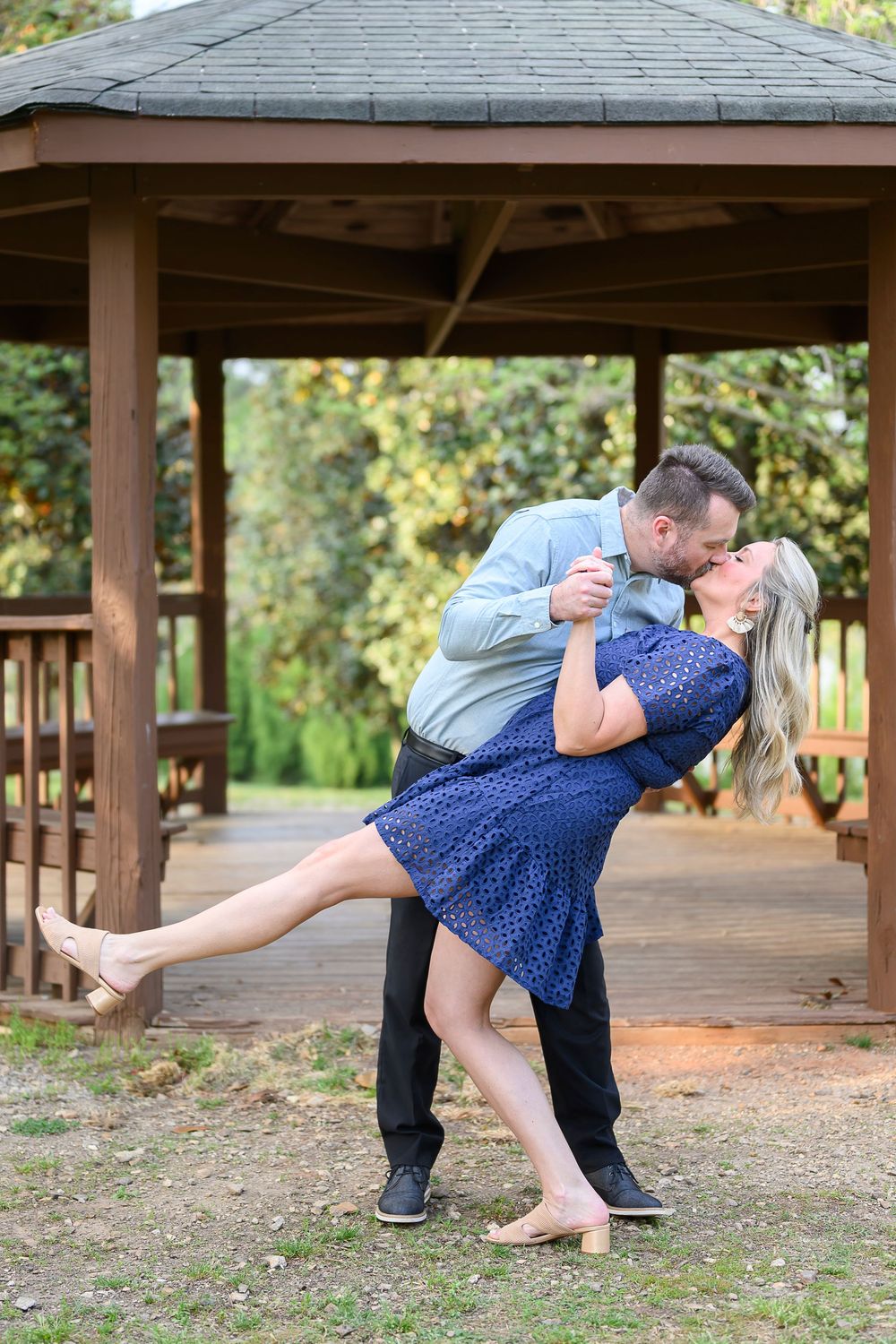 Couple sharing a romantic dip kiss in front of a wooden gazebo, woman in navy dress, man in blue shirt.
