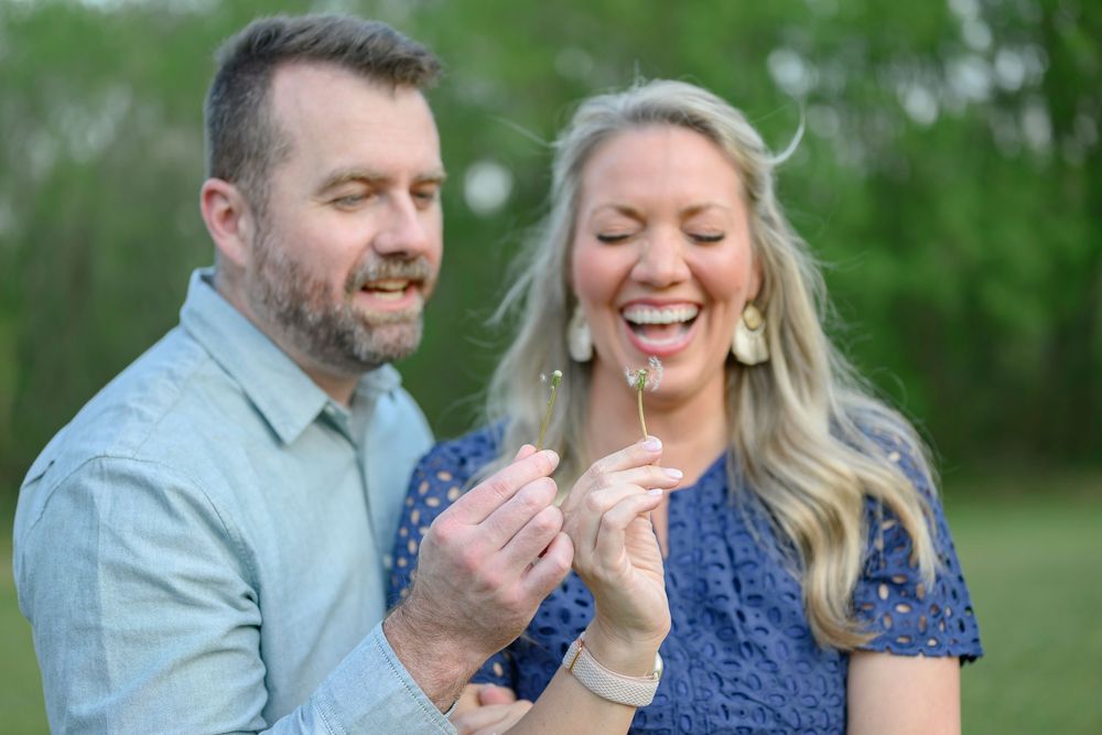 Couple laughing and blowing a dandelion together outdoors in a green field during a romantic engagement photo session.