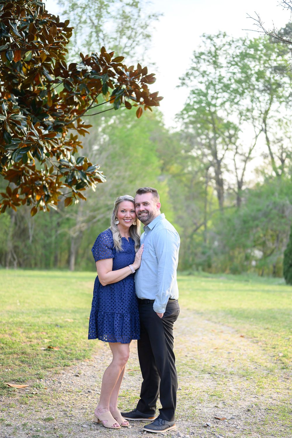 A smiling couple poses together on a garden path surrounded by lush green trees and magnolia leaves.
