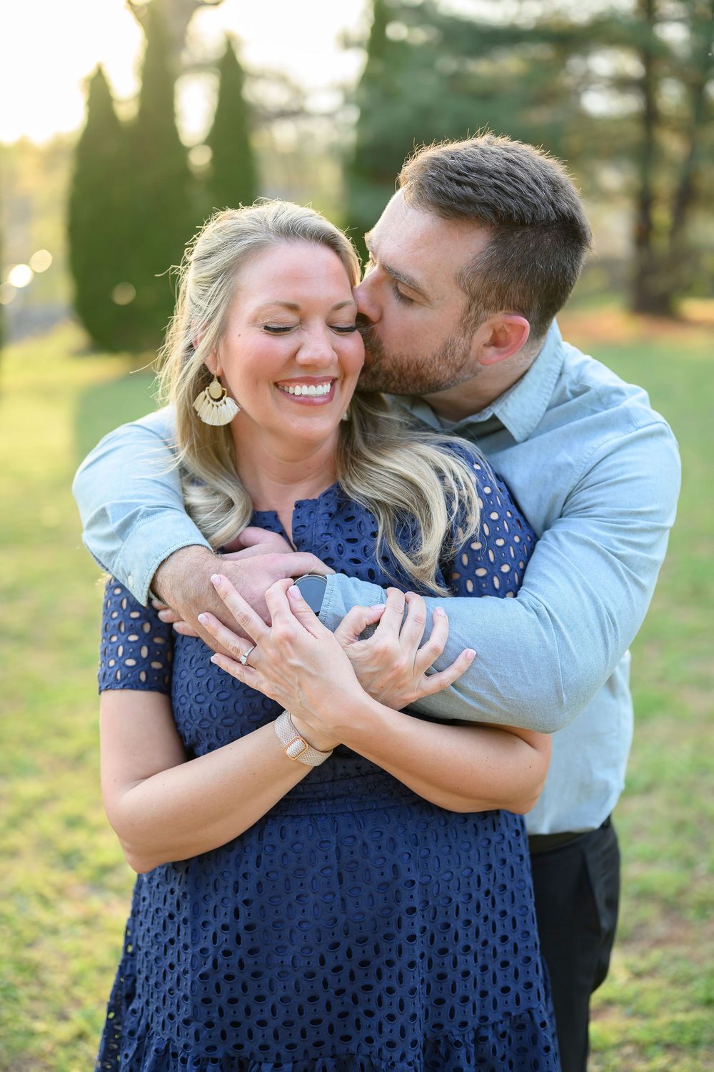 A smiling woman in a navy dress is embraced and kissed on the cheek by a man in a light blue shirt outdoors.