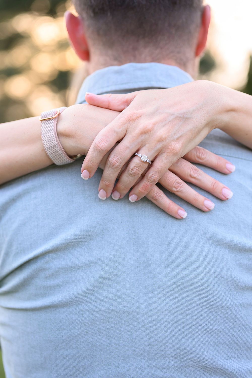 Woman showing diamond engagement ring while embracing man from behind, wearing pink mesh bracelet watch.