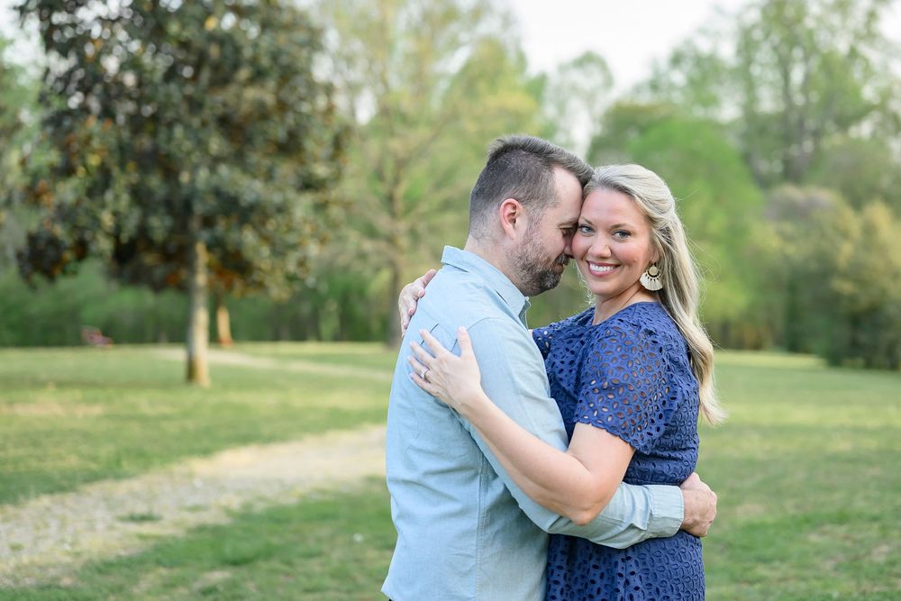 Couple embracing and smiling in a lush green park, woman in blue lace dress, man in light blue shirt.