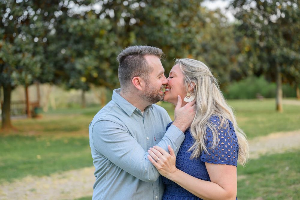 A couple shares a romantic kiss outdoors in a lush green park, the man in blue and woman in a lace dress.