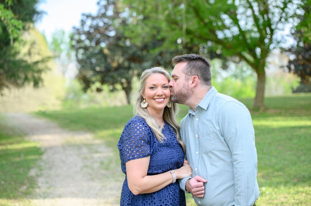 Couple sharing a tender moment outdoors, man kissing woman's cheek on a scenic green park path.