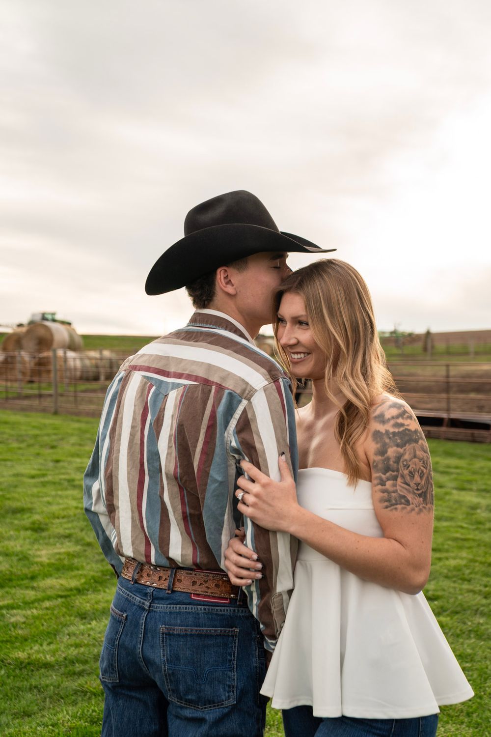 A couple in western attire embraces outdoors, the man in a black cowboy hat kissing her cheek as she smiles on a farm.