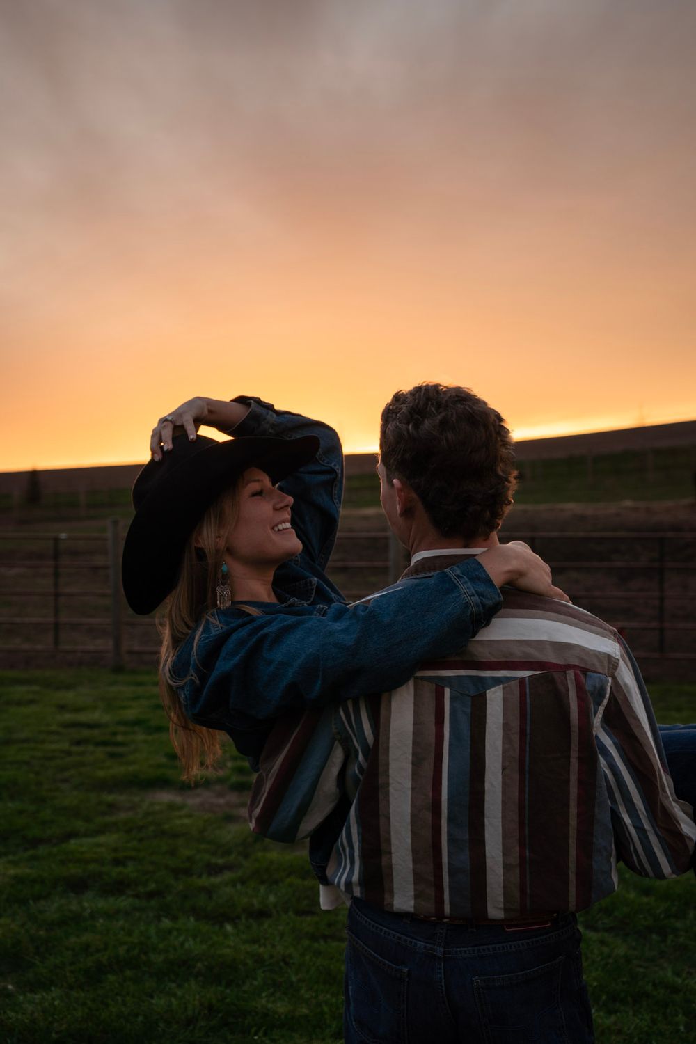 Woman smiling and placing black cowboy hat on her head while embracing man at golden sunset on ranch.