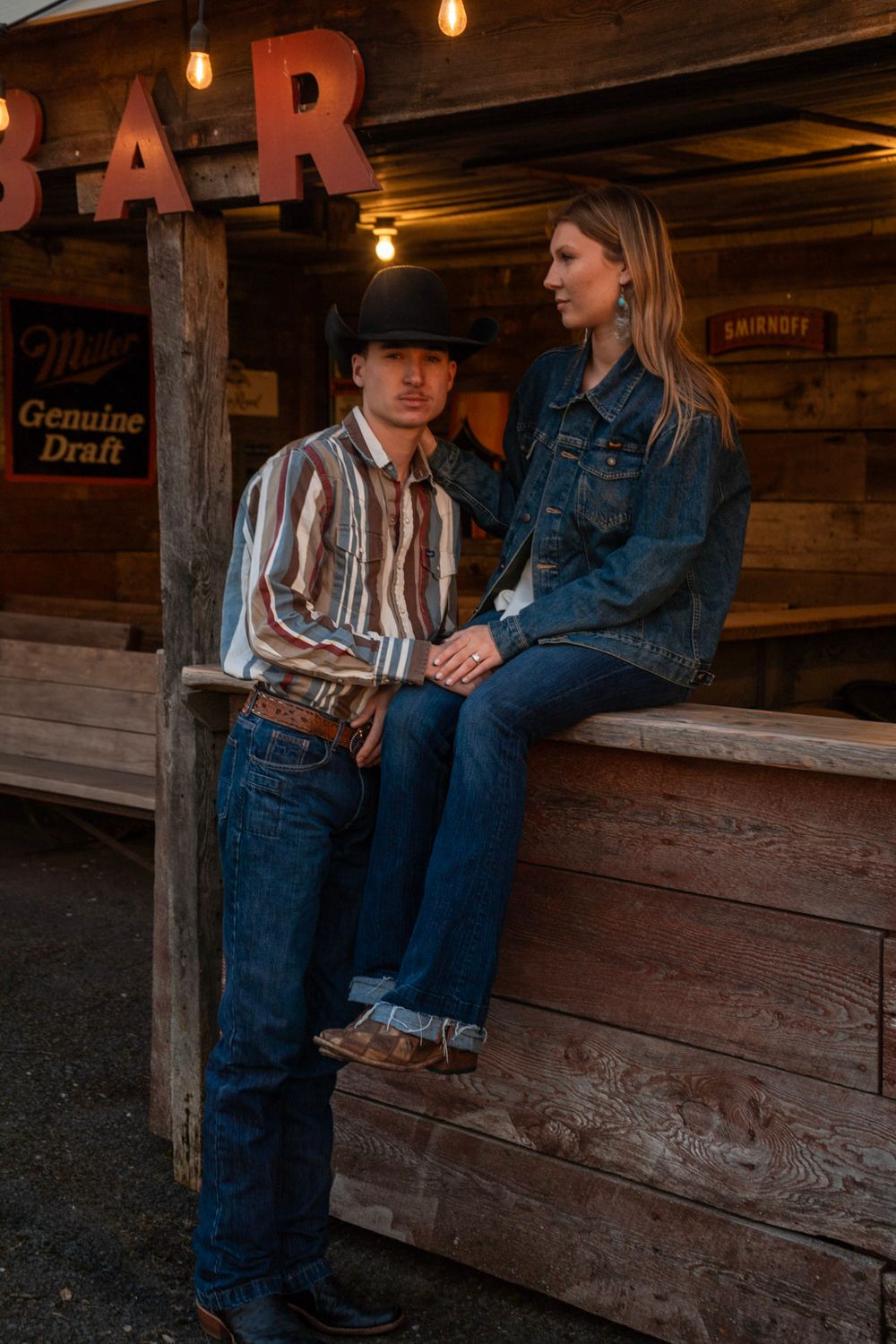 Couple in western wear posing outside a rustic bar, man in cowboy hat and striped shirt, woman in denim jacket.