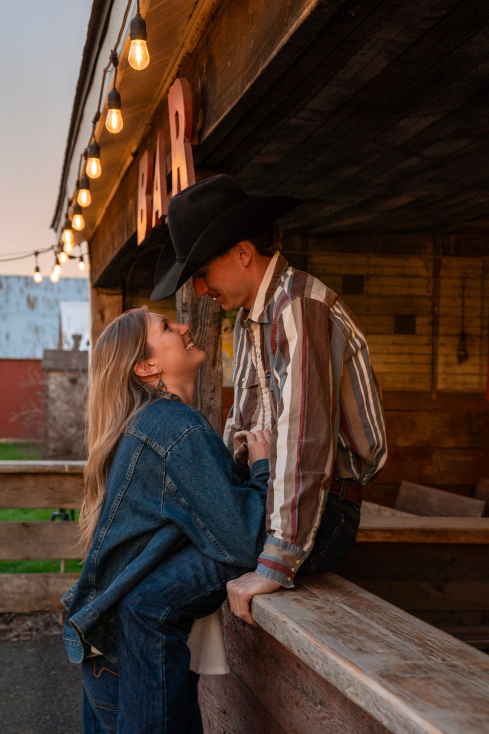Romantic couple nearly kissing outside a rustic bar at dusk, man in cowboy hat and striped western shirt.