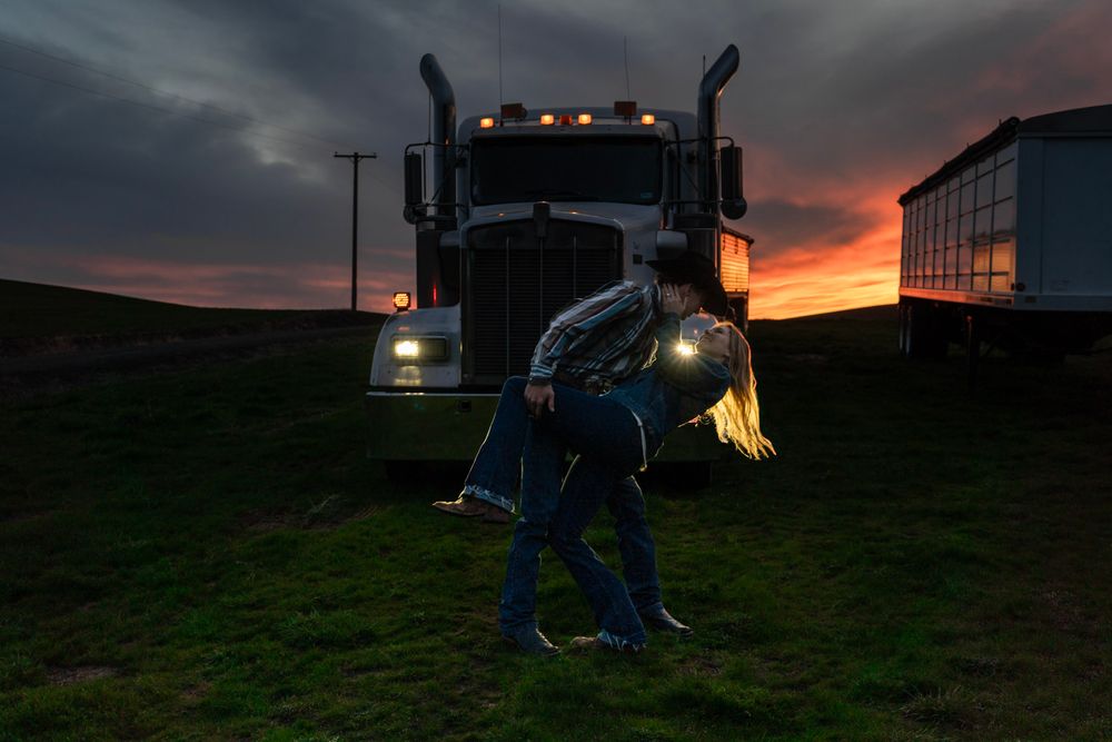 Couple sharing a romantic dip kiss at sunset in front of a semi-truck on a rural highway.