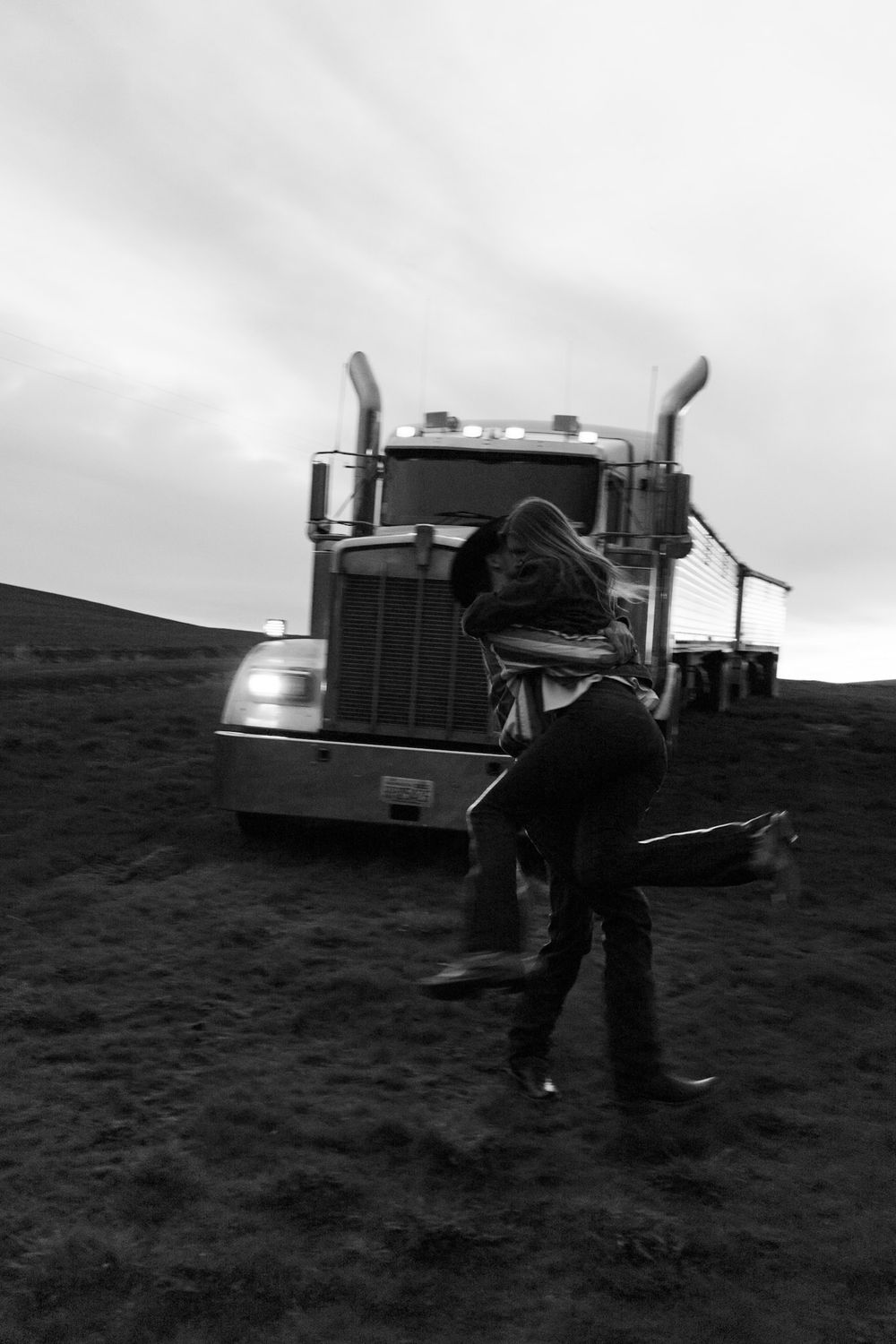 Black and white photo of couple embracing in front of a semi truck on a rural farm field under cloudy skies.