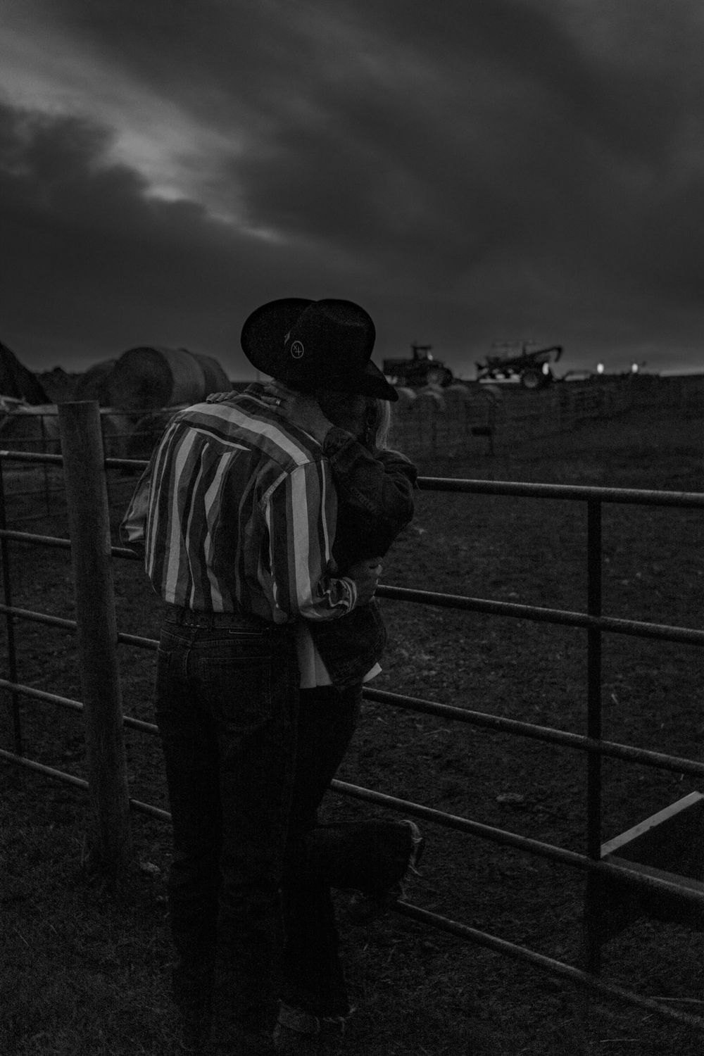 Black and white photo of a cowboy in striped shirt and hat leaning on a metal fence gate at dusk on a farm.