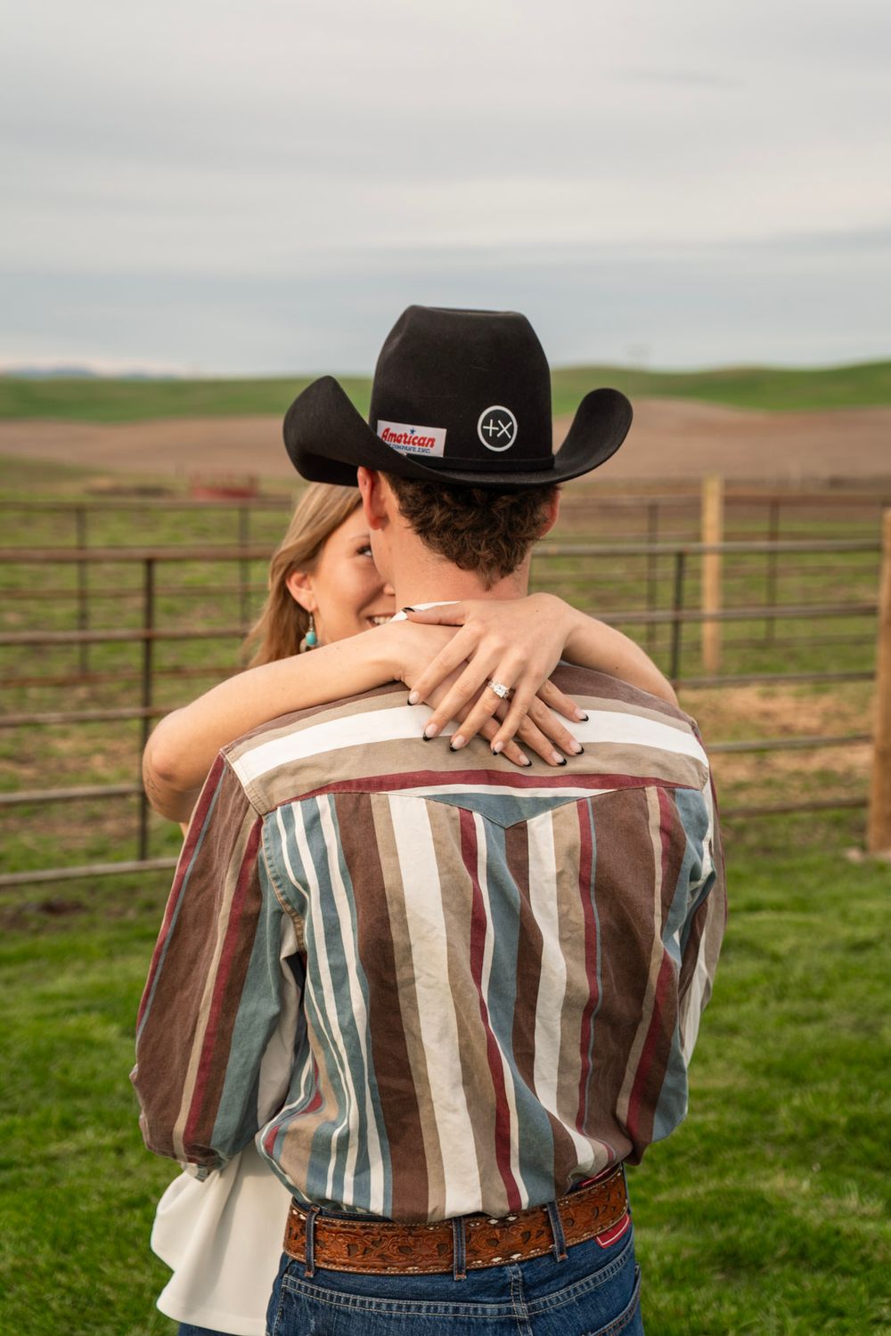 Couple embracing at a ranch, man in black cowboy hat and striped western shirt, woman showing engagement ring.