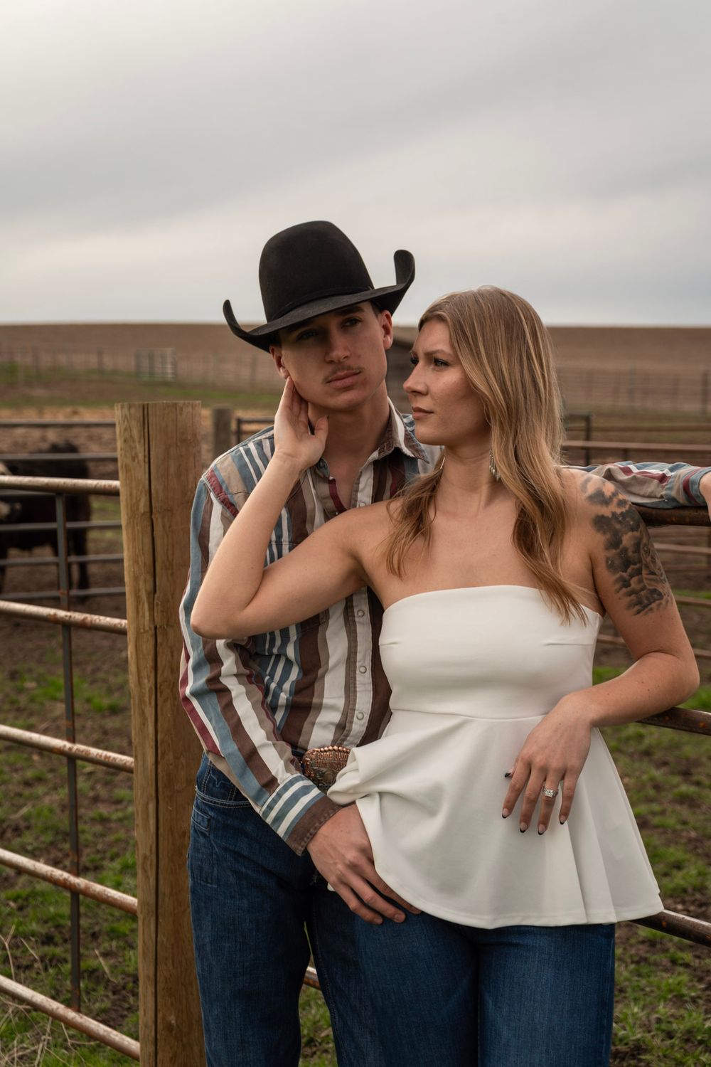 A couple poses by a ranch gate, man in cowboy hat and plaid shirt, woman in white strapless top with tattoo.