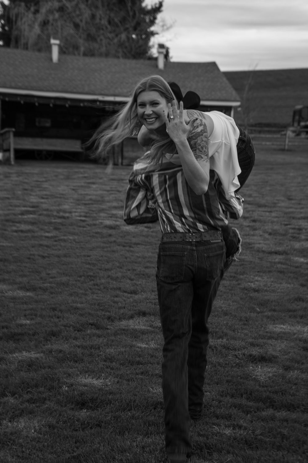 Smiling blonde woman carrying someone on her back in a field near a barn, black and white photo.