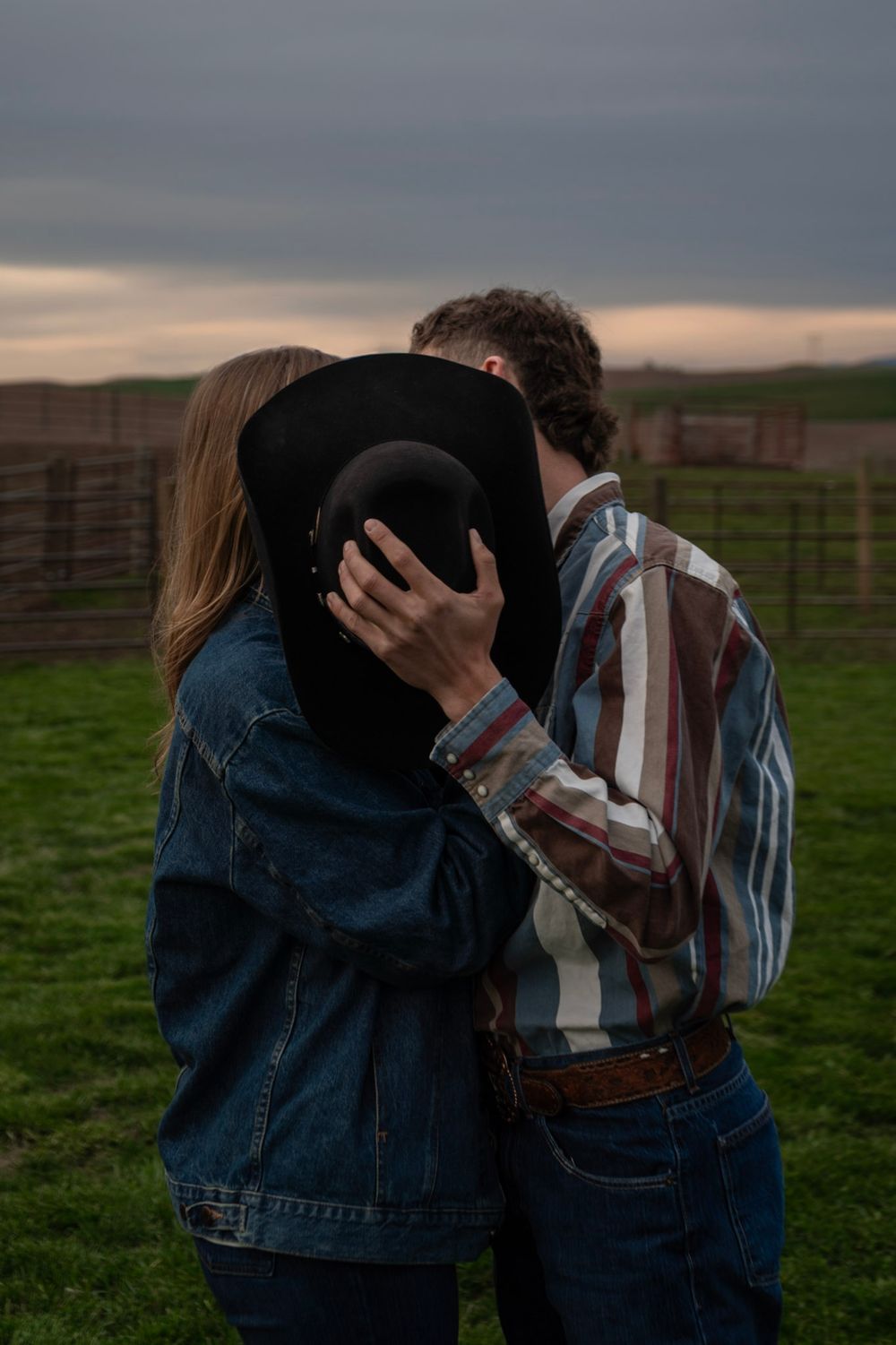 Couple sharing a kiss hidden behind a black cowboy hat at sunset on a rural farm.