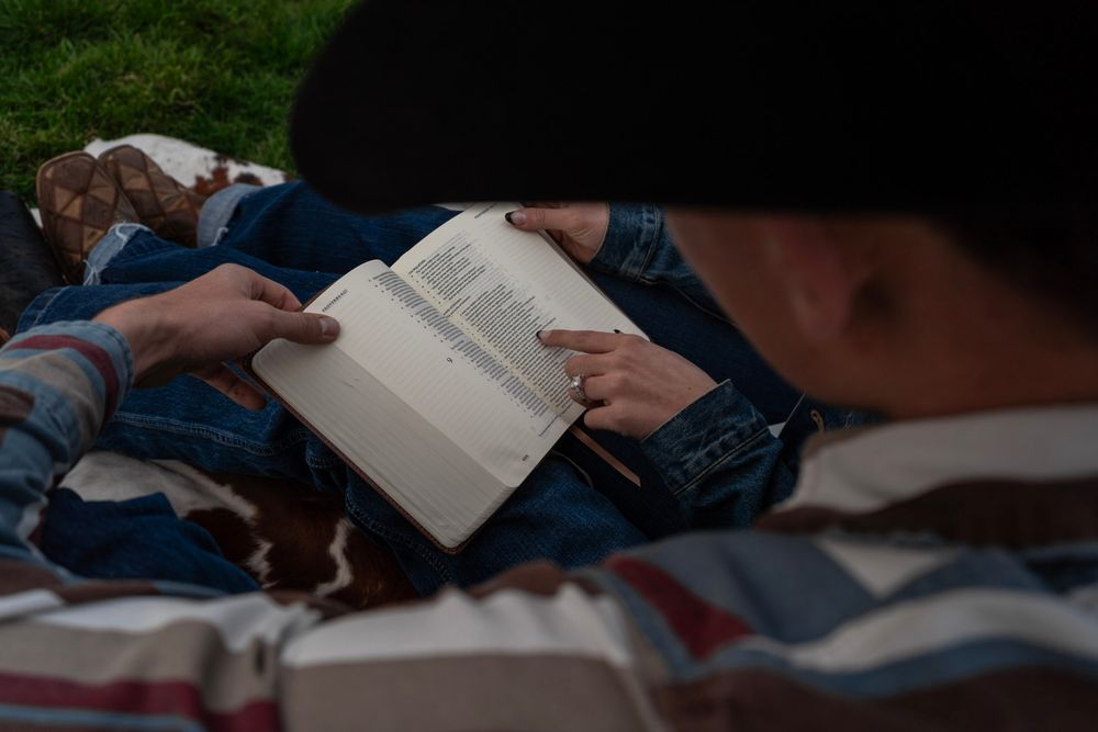 Two people lying on grass reading a book together outdoors on a sunny day.