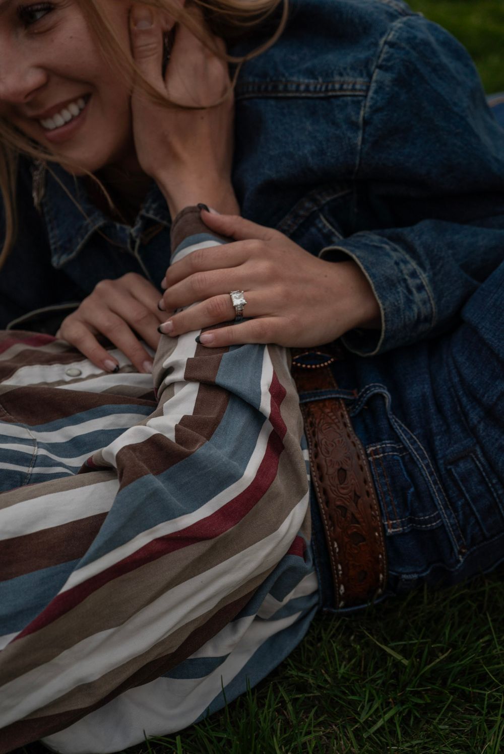 Close-up of smiling woman wearing engagement ring hugging man in striped western shirt on grass.