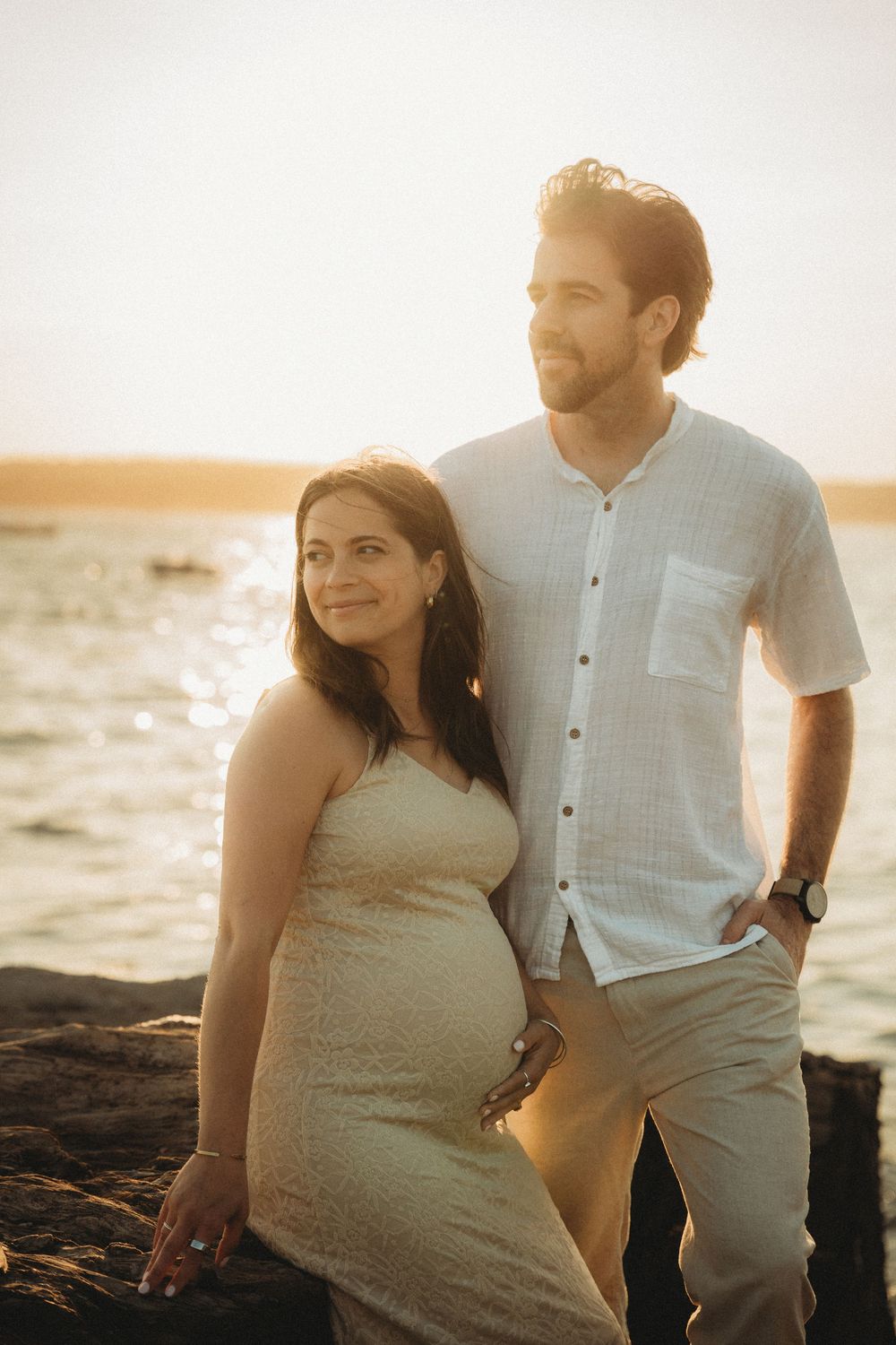 Pregnant couple posing at golden hour by the ocean, woman in white dress with baby bump, man in light blue shirt smiling.