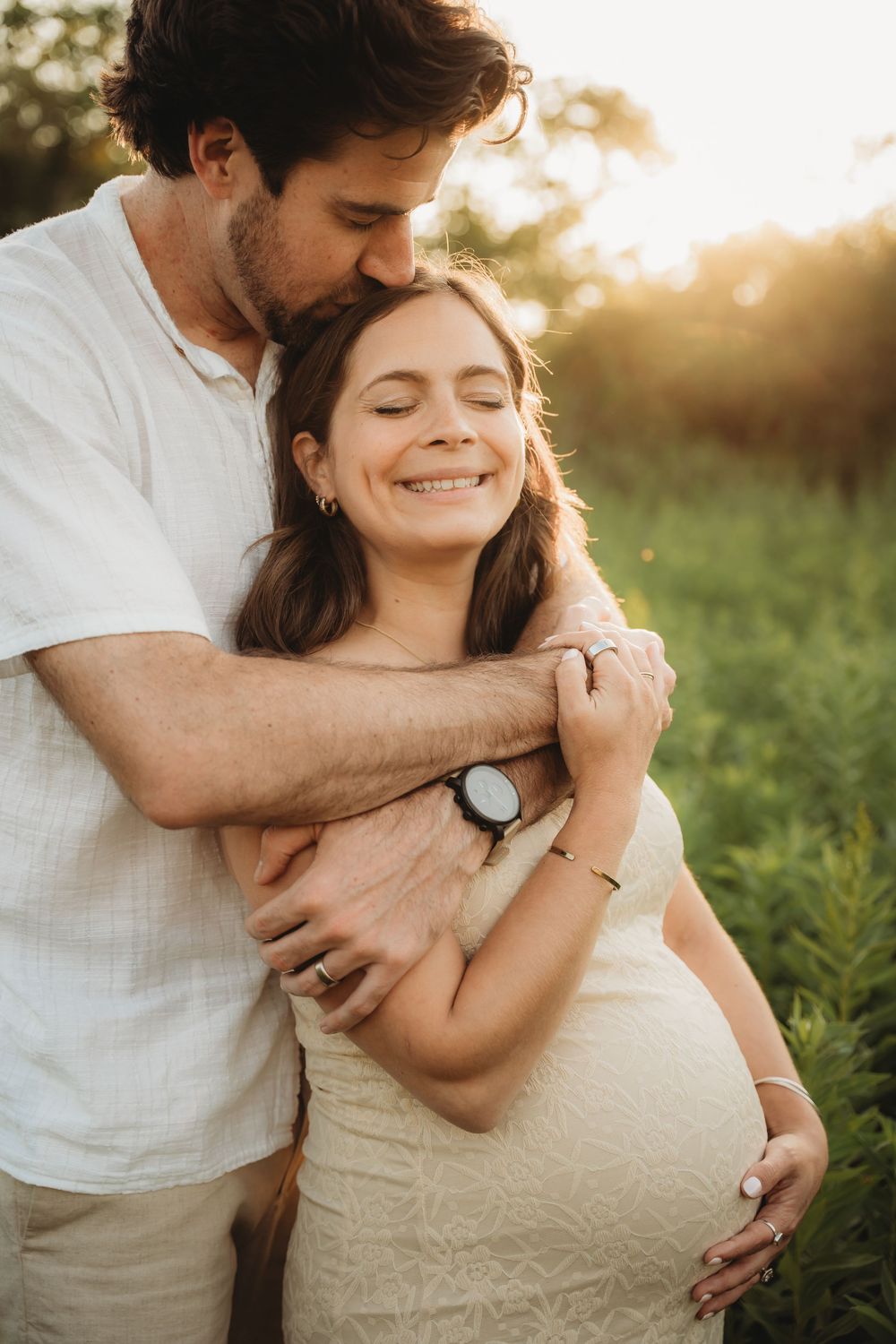 A man tenderly embraces and kisses a smiling pregnant woman in a sunlit green field during golden hour.