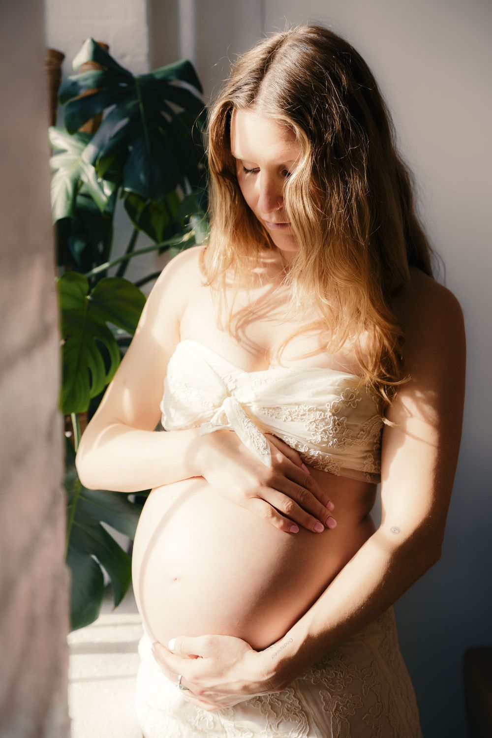 Pregnant woman in white lace bra cradling her bare baby bump, standing by a window with monstera plant.