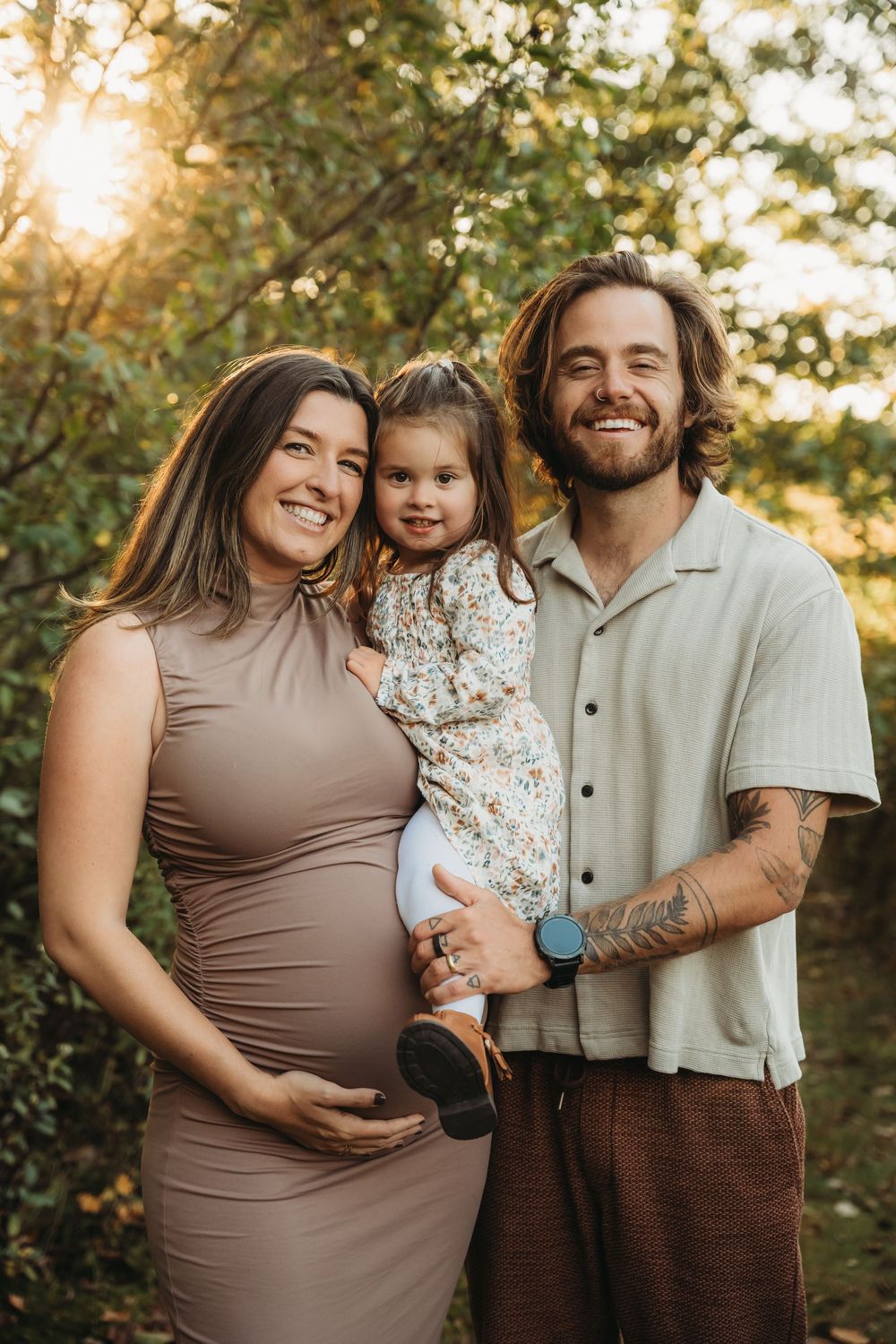 Pregnant woman in beige dress poses with smiling man and toddler girl outdoors in golden sunlight.