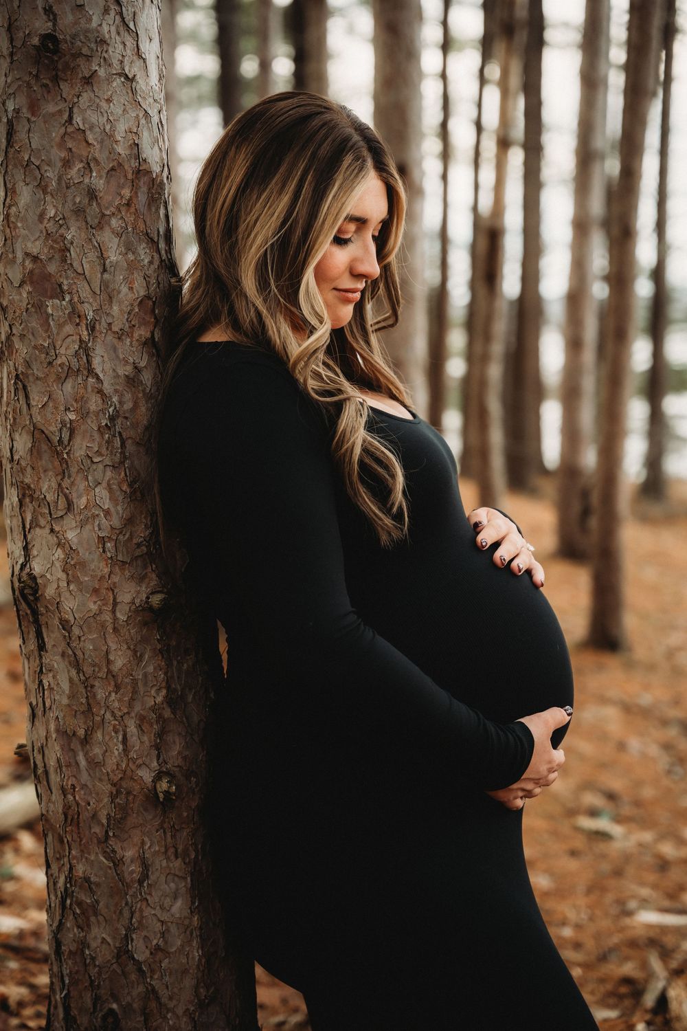 Pregnant woman in black dress leaning against tree in forest, cradling baby bump during outdoor maternity photo session.