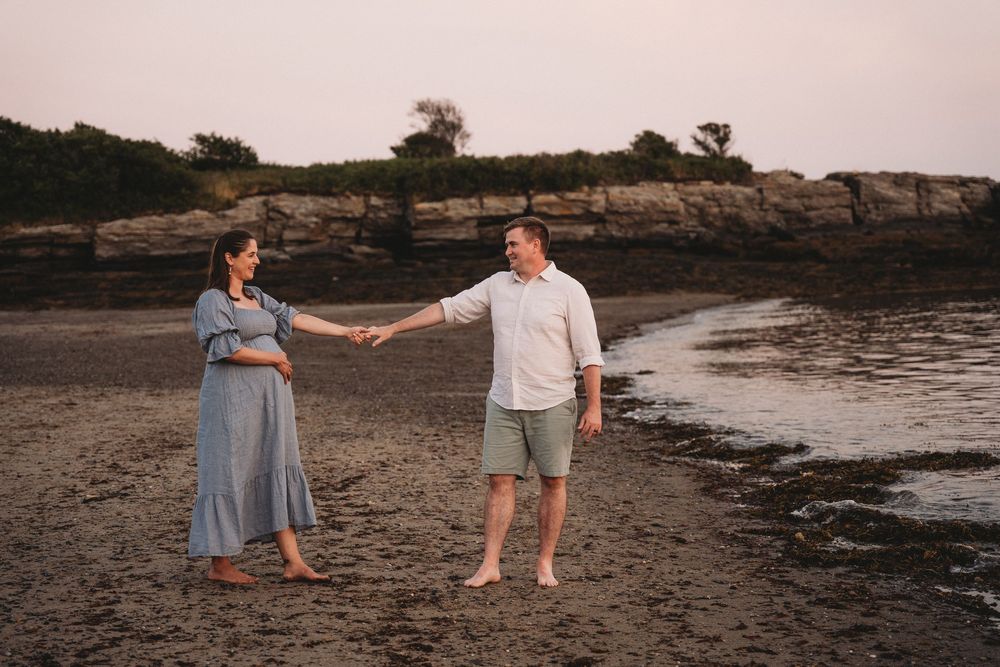 Pregnant couple holding hands on rocky beach at sunset, woman in blue dress, man in white shirt and green shorts.