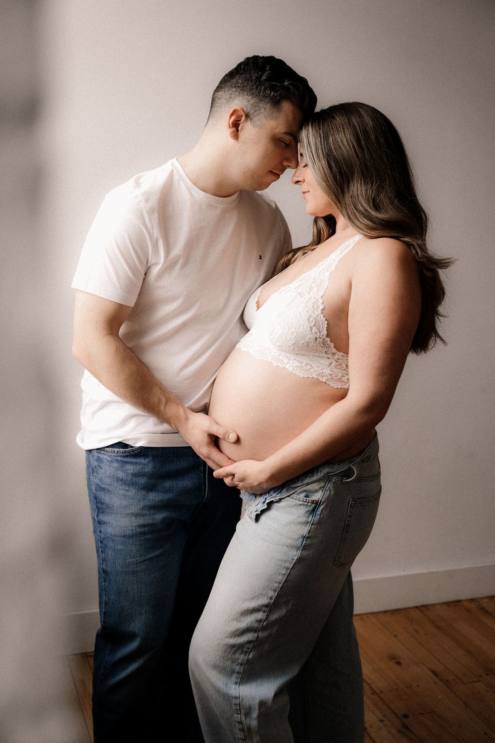 Couple sharing tender moment during maternity photo shoot, man in white tee embracing pregnant woman in white lace bralette.