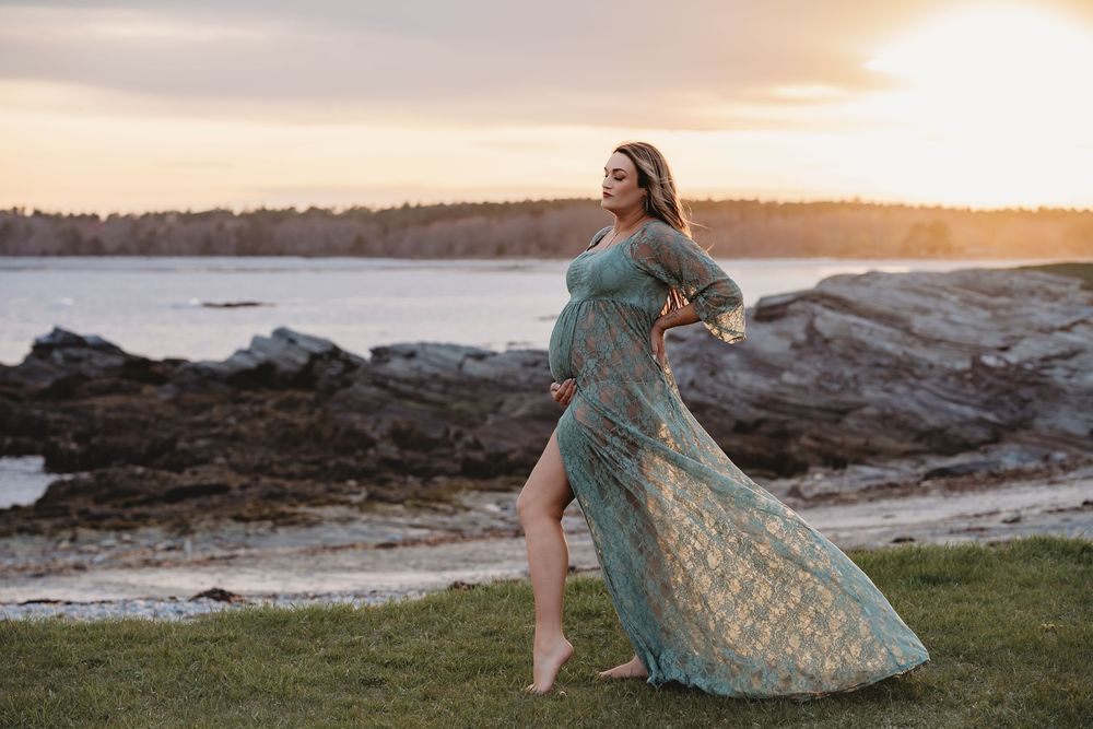 Pregnant woman in teal lace gown poses on rocky coastal shore at golden sunset.