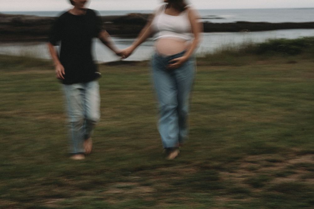 Pregnant couple holding hands walking outdoors near water in casual jeans attire.