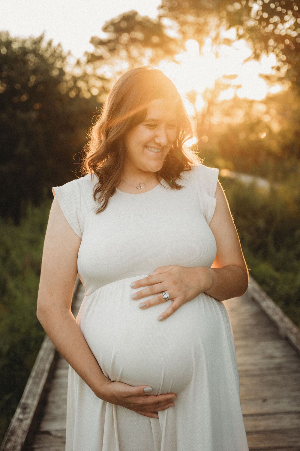 Smiling pregnant woman in white dress cradling baby bump at golden hour on a wooden boardwalk.