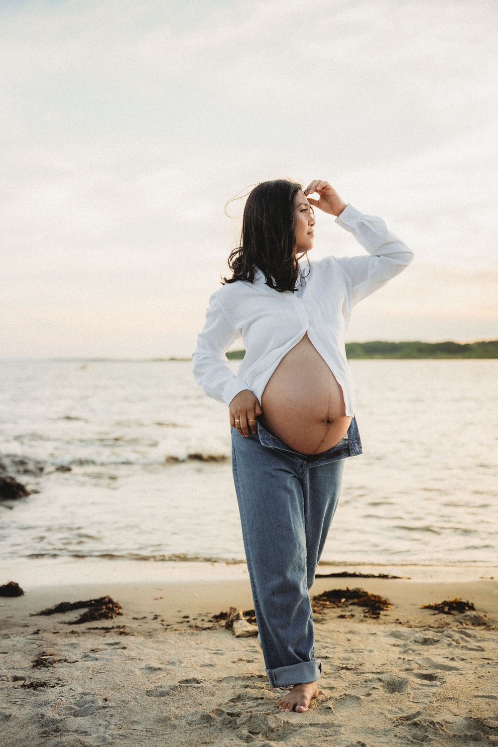 Pregnant woman in white shirt and jeans standing on a sandy beach at sunset, showing her baby bump. Maine