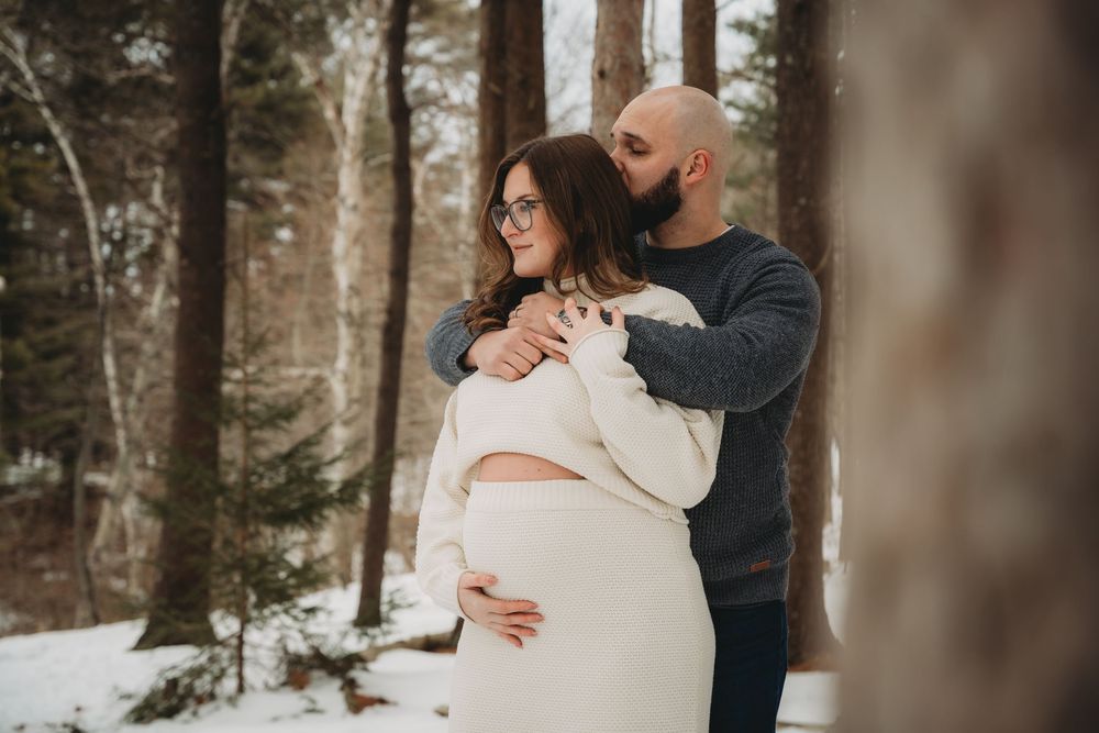 Couple embracing in snowy winter forest, pregnant woman in white dress held by partner among snow-covered pine trees.