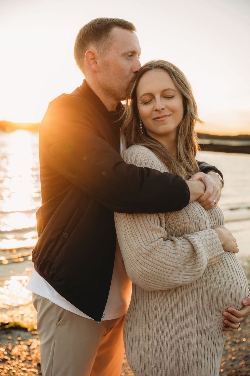 Couple embracing at golden hour sunset on rocky beach, man kissing pregnant woman's temple in tender maternity photo.