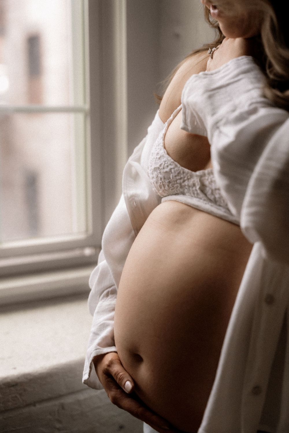 Pregnant woman in white lace bra and open shirt cradling her bare baby bump by a sunlit window.