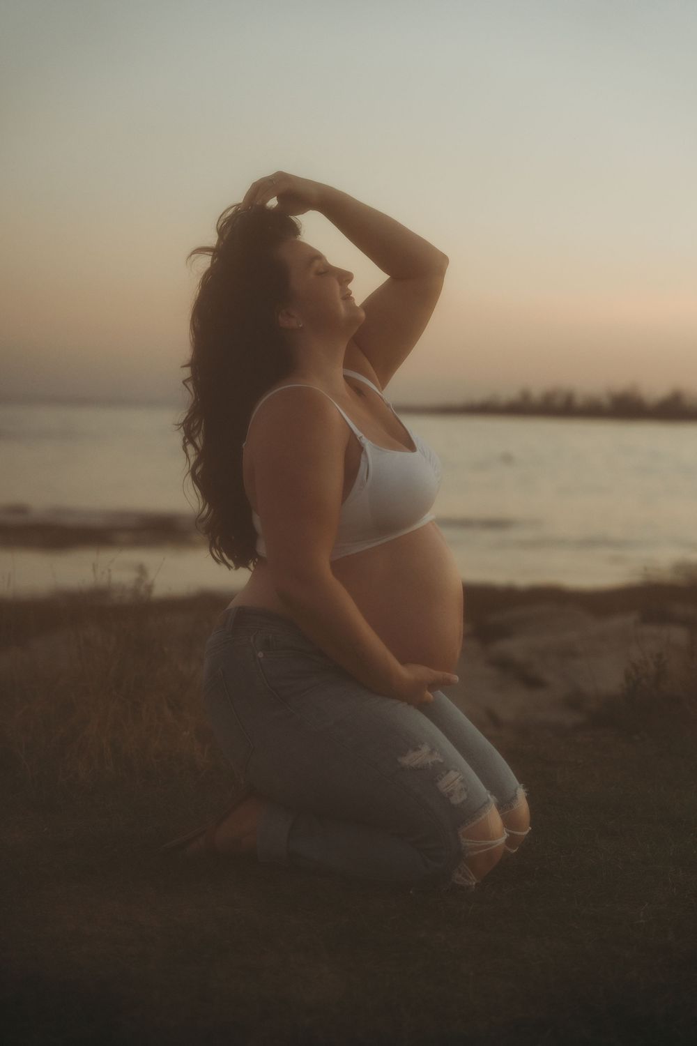 Pregnant woman in white crop top and jeans kneeling outdoors at sunset near water, posing with hand in hair.