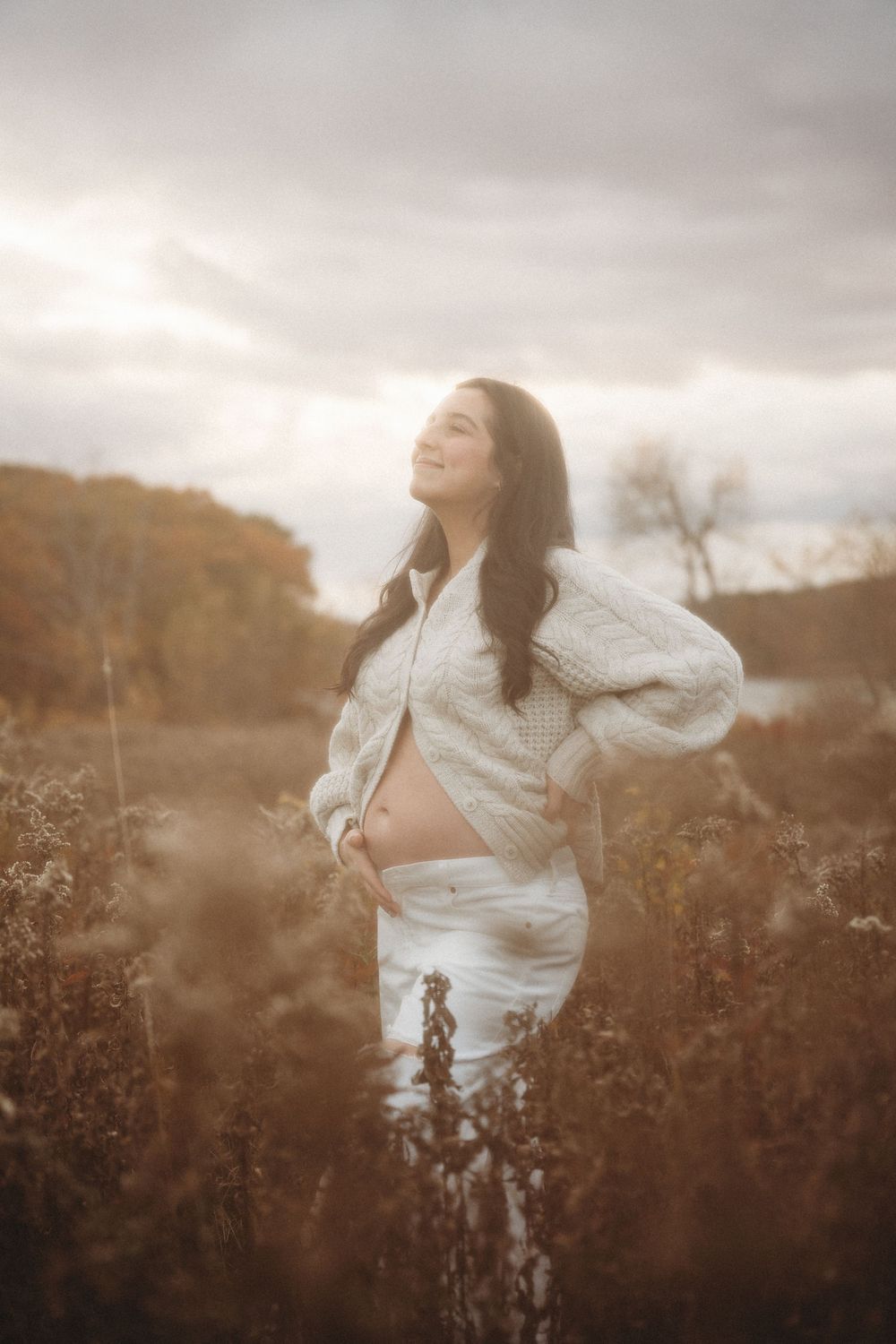 Smiling pregnant woman in white outfit standing in autumn field, cradling baby bump, with golden foliage and cloudy sky.