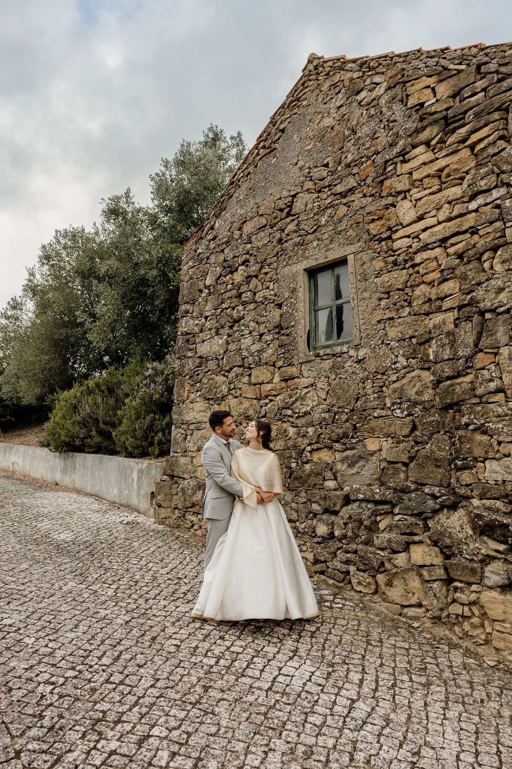 A couple in wedding attire embraces against a rustic stone wall on a cobblestone path.