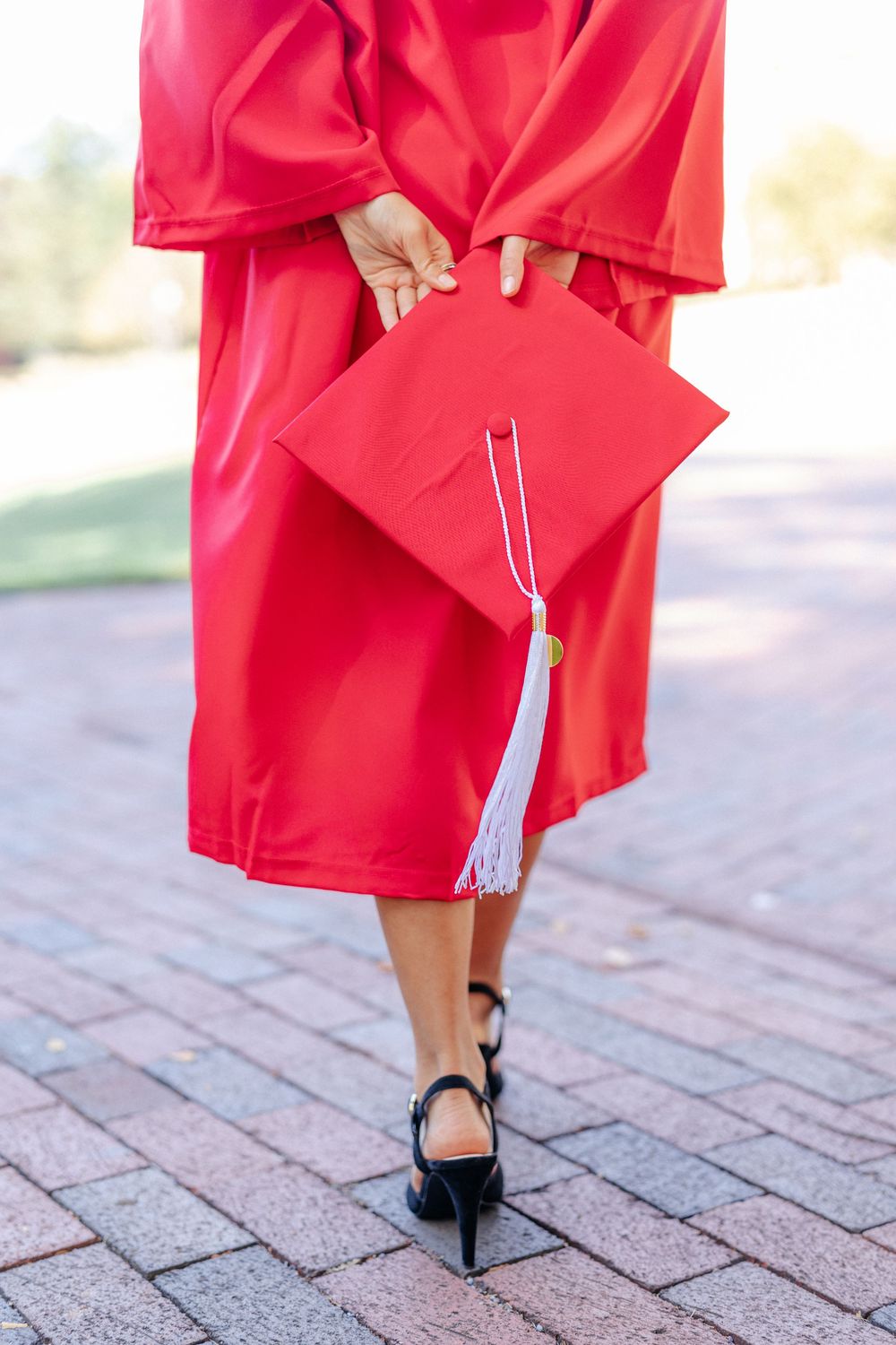 Graduate in red cap and gown walking away holding mortarboard cap on a brick pathway outdoors.
