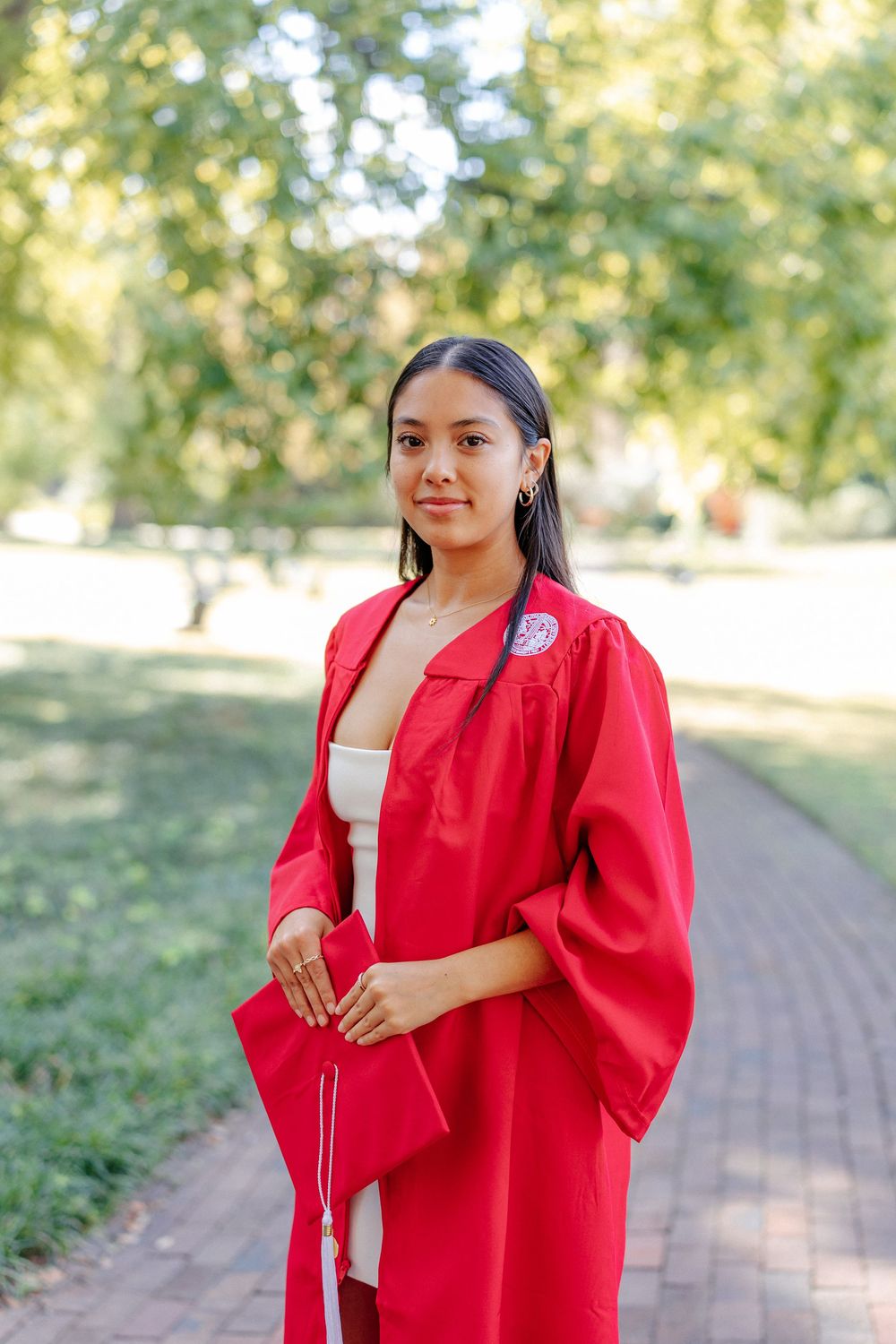Young woman in red graduation gown and cap standing on a tree-lined brick pathway for graduation portraits.