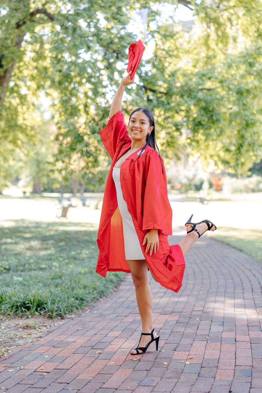Smiling woman in red graduation gown raises cap while posing on brick path in sunny park.