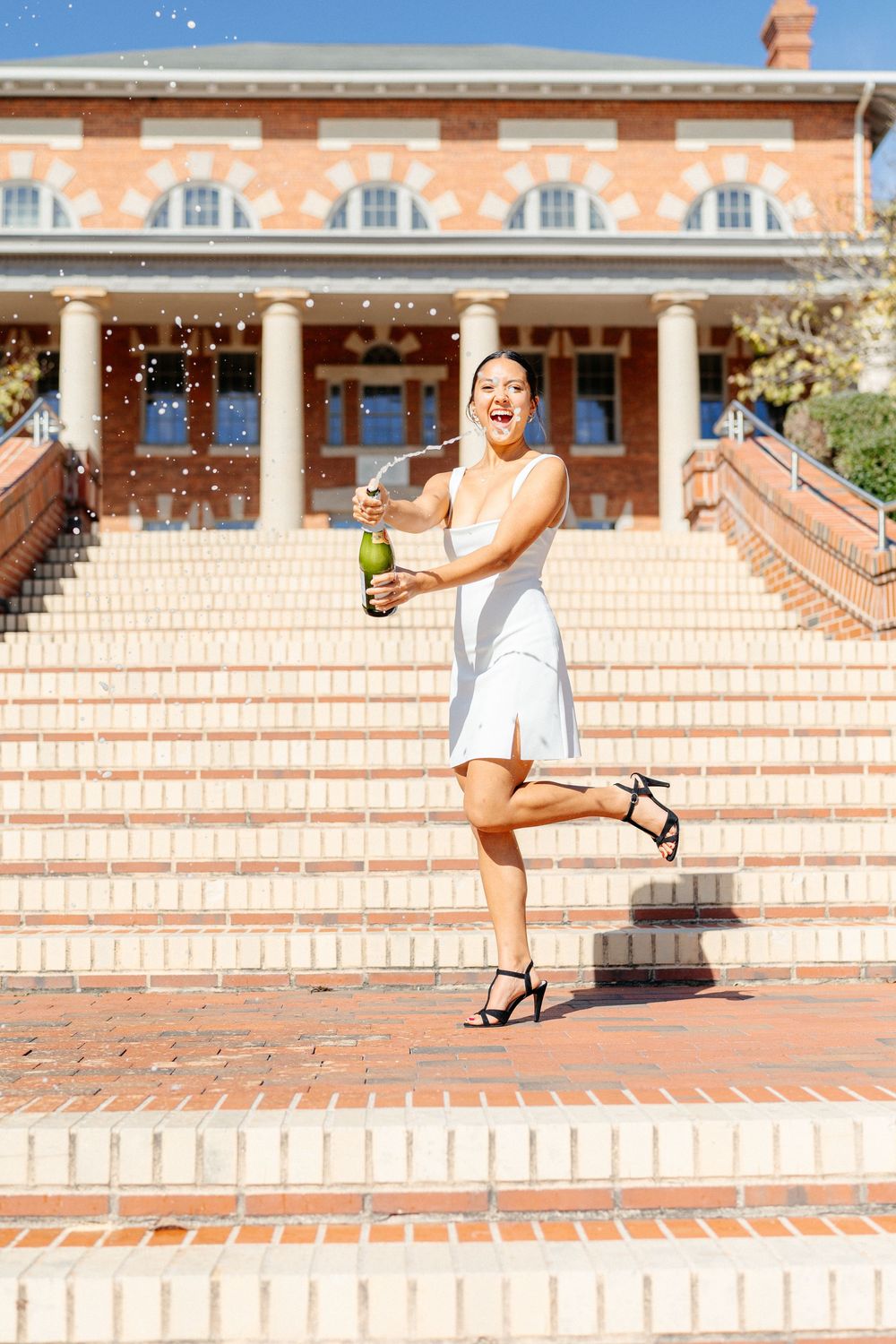 Excited woman in white dress pops champagne bottle on steps outside brick building.