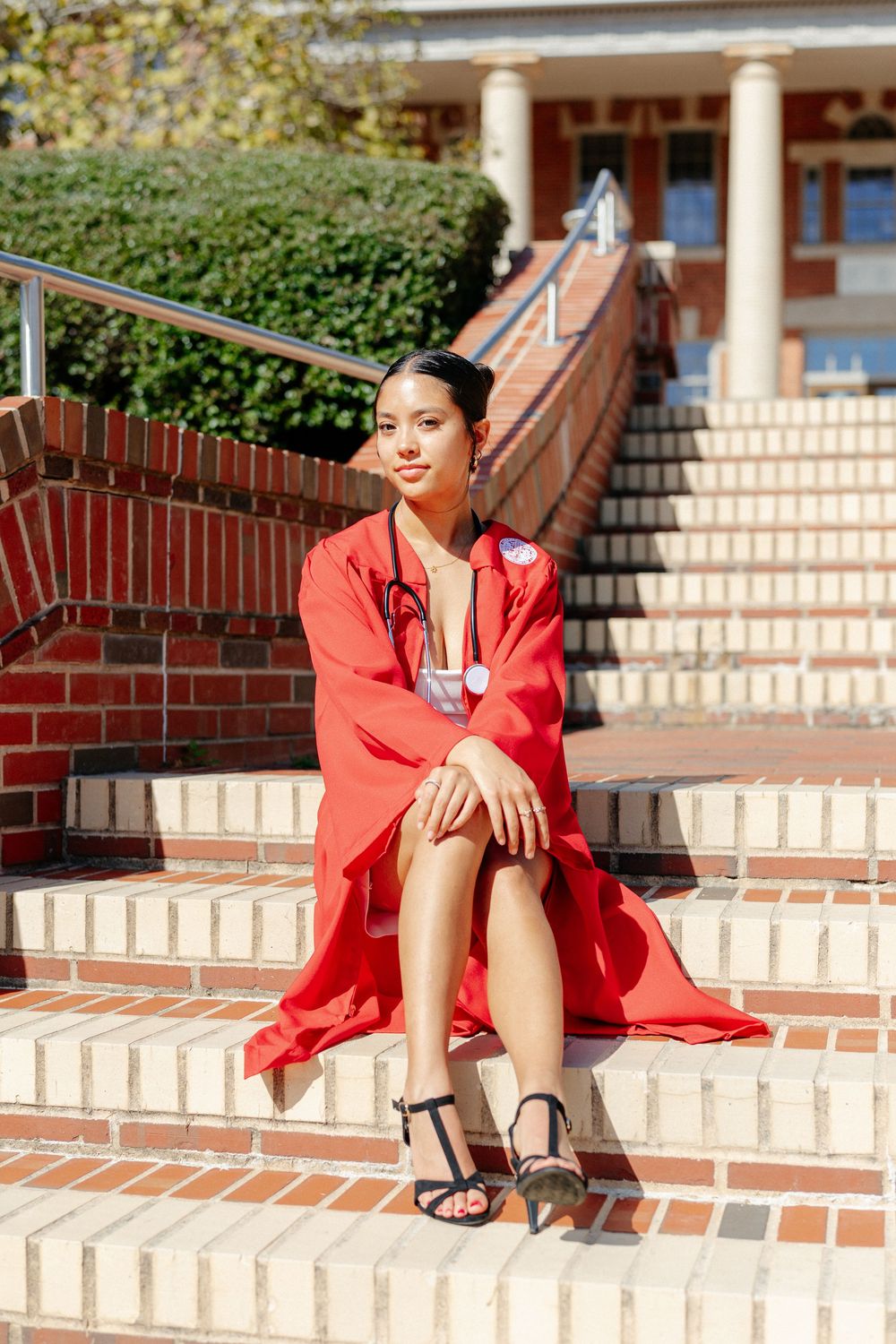 Young woman in red graduation gown with stethoscope sits on brick steps outdoors.
