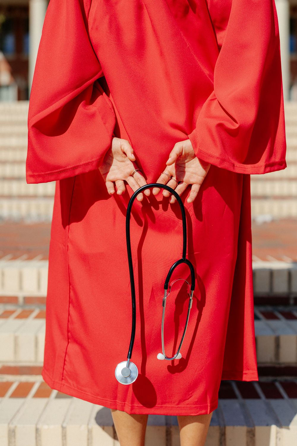 Close-up of person in red graduation gown holding stethoscope behind their back.