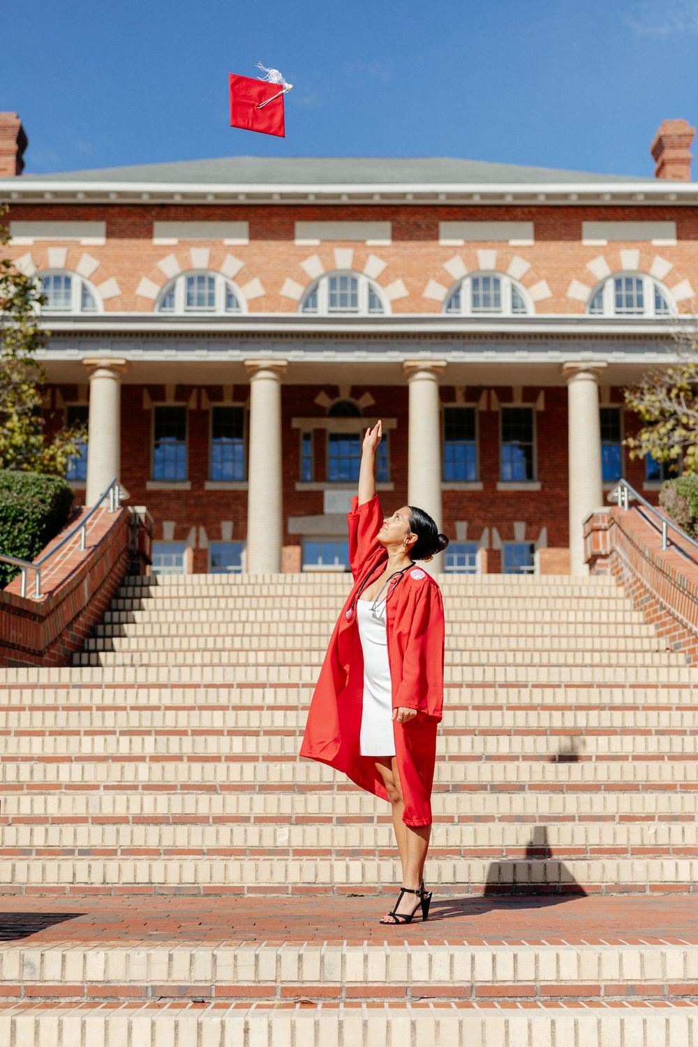Woman in red graduation gown tosses cap in air in front of columned brick building.